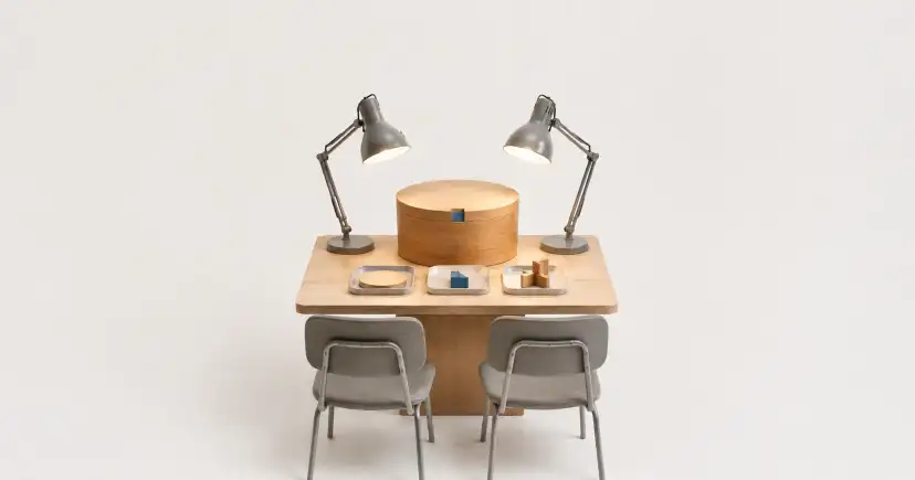 Mesa de trabajo para dos personas, con lámparas, sillas y varias piezas de madera organizadas.