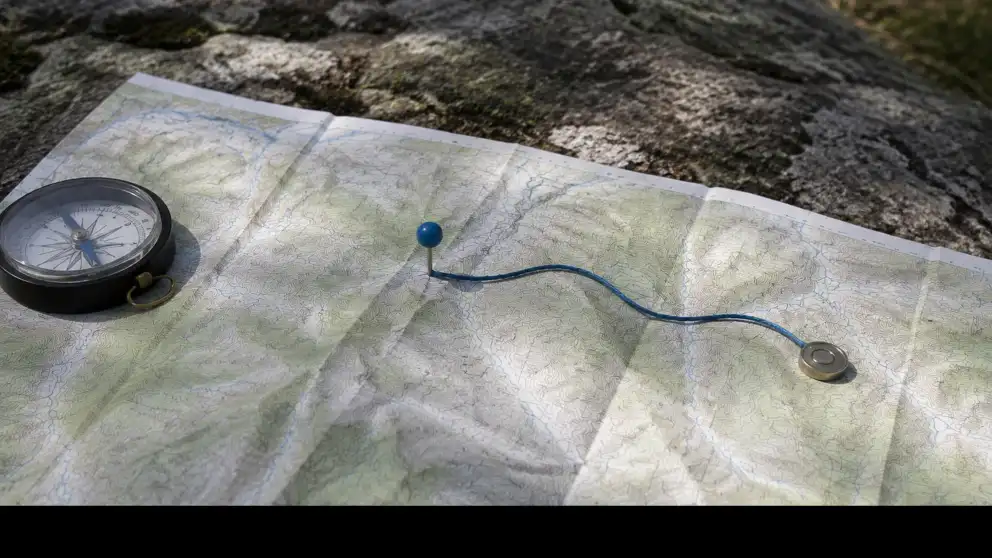 Mapa topográfico con una brújula y una ruta azul trazada entre un alfiler y un marcador metálico.