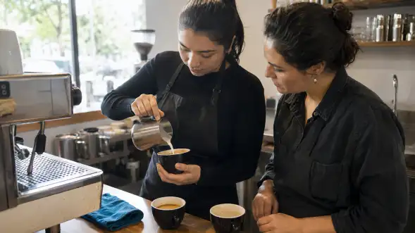Barista vierte leche en una taza de café mientras una compañera observa.