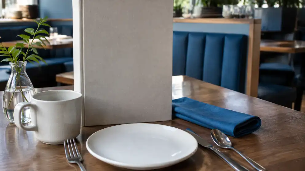 Mesa de cafetería con un plato, una taza, cubiertos, una servilleta azul y un menú en blanco.