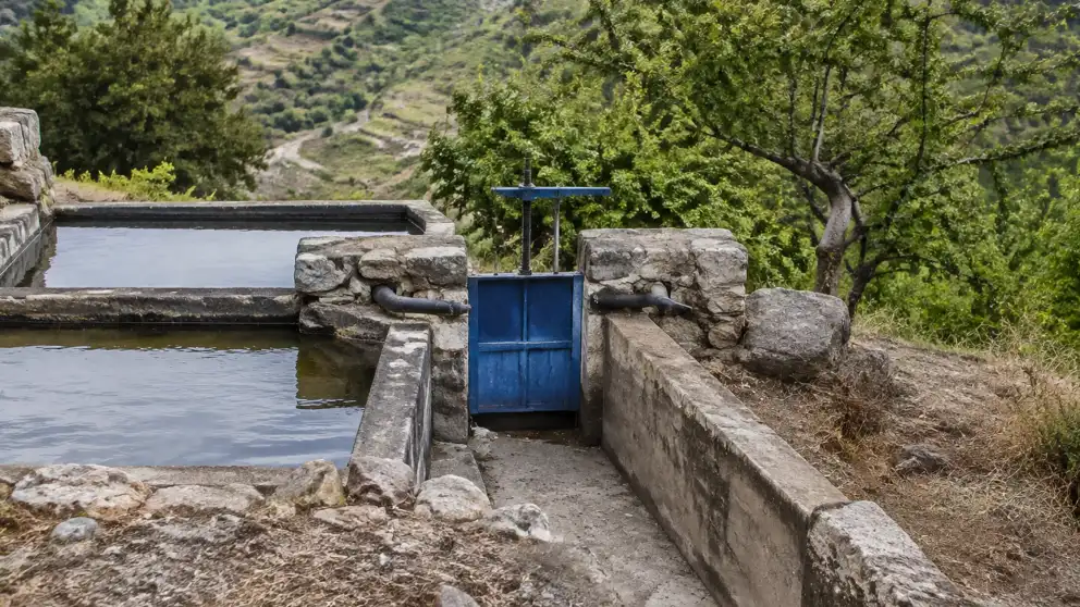 Una compuerta azul regula el agua que circula por un canal de riego de piedra en la montaña.
