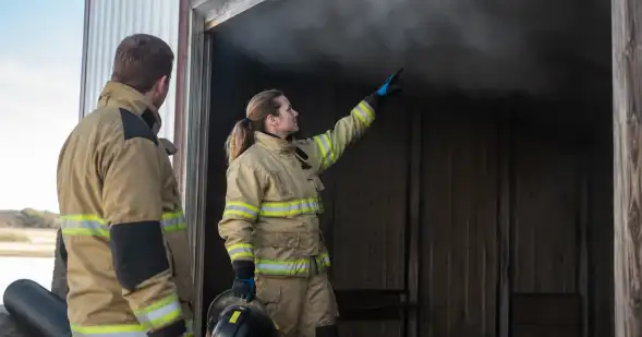 Dos bomberos inspeccionan el interior ennegrecido de una estructura y señalan marcas sobre la pared.