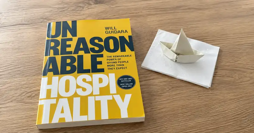 Portada amarilla de Unreasonable Hospitality sobre una mesa de madera, junto a un barco de papel blanco.