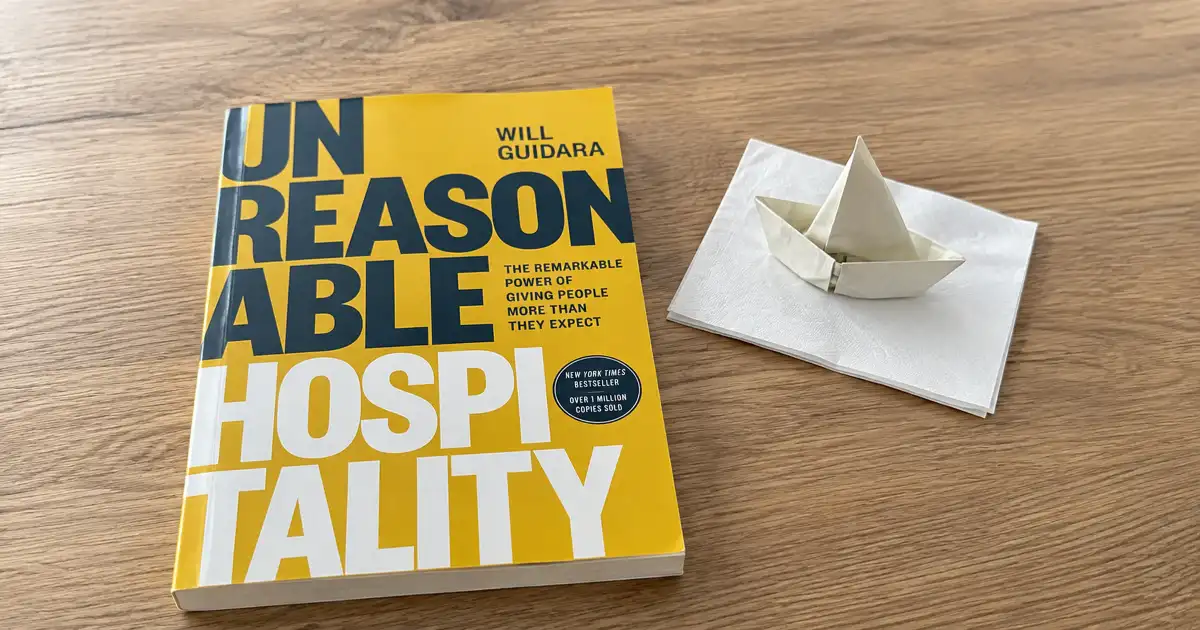 Portada amarilla de Unreasonable Hospitality sobre una mesa de madera, junto a un barco de papel blanco.