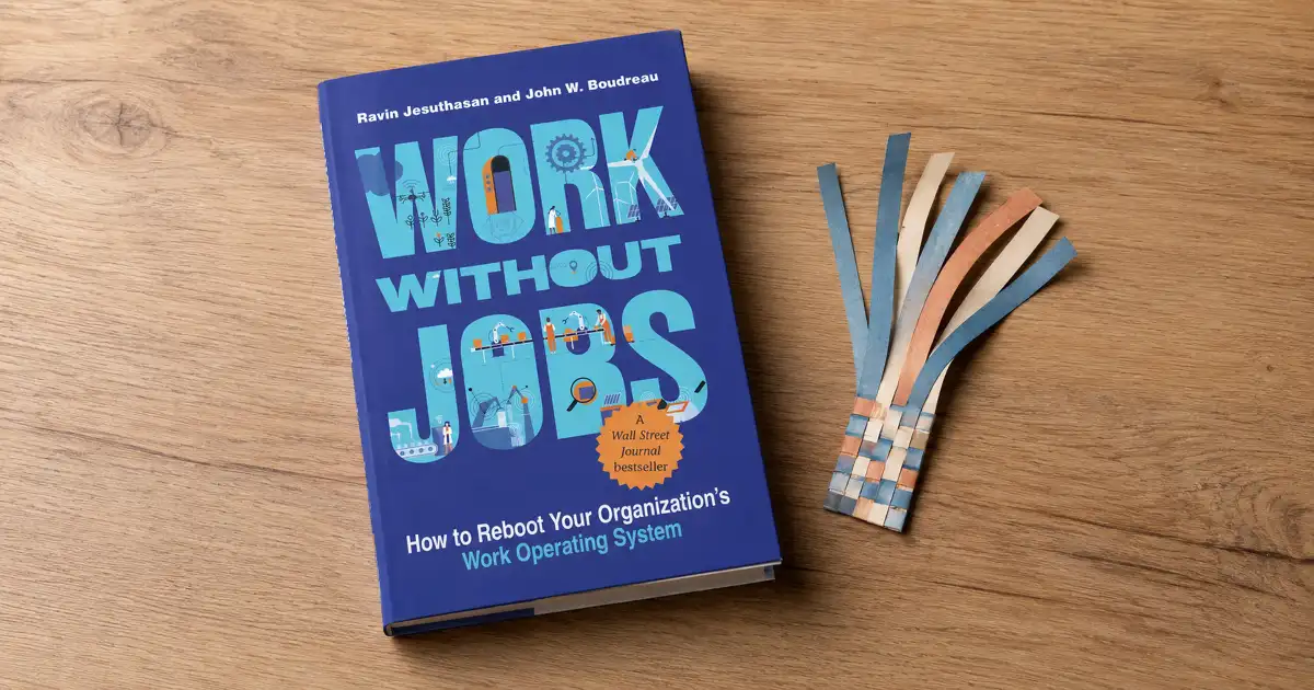 Portada de Work Without Jobs sobre una mesa de madera, junto a tiras de papel multicolores entrelazadas.